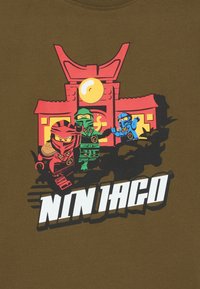 Olivegrünes T-Shirt mit einem bunten Grafikdruck von drei Ninja-Figuren in Rot, Grün und Blau, vor einem stilisierten Tempel-Hintergrund und weißem Text.