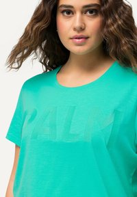 Camiseta verde de manga corta hecha de tela suave, con letras texturizadas "PALM" en un tono más claro. Cuello redondo clásico.
