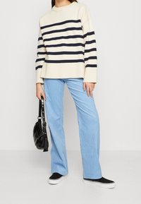 Cremfarbener und marineblauer gestreifter Strickpullover mit lässigem Schnitt, kombiniert mit hellblauen Baumwollcord-Hose im weiten Schnitt und schwarzen Slippern.