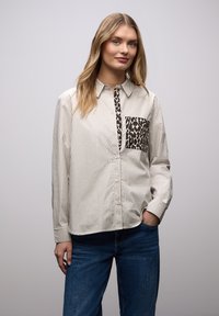 Chemise à boutons rayée beige et blanche, avec une poche à imprimé léopard. Le tissu semble lisse, avec une coupe décontractée et des manches longues.