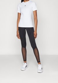 Kvinna iklädd vit Adidas kortärmad tröja, svarta tights med nätpaneler och vita Adidas sneakers, stående mot en enkel bakgrund.