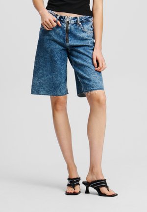 Personne portant un short en jean bleu mi-long avec fermeture éclair à l'avant et des sandales noires à talons avec des sangles, debout devant un fond uni.