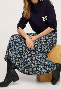 Sweat-shirt bleu marine avec un écusson blanc en forme de "M", longue jupe noire à fleurs avec des motifs bleus, roses et verts, et bottes en cuir noires montant jusqu'aux genoux.