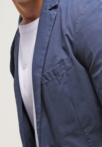 Námořnický modrý, lehký blazer s hladkým povrchem, s klopou do písmene V a jednou náprsní kapsou, nošený přes bílé tričko s kulatým výstřihem.