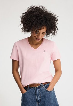 Kobieta o kręconych włosach, ubrana w jasnoróżowy T-shirt z dekoltem w serek i niebieskie dżinsy z brązowym paskiem, ręce w kieszeniach, patrząca w dół.