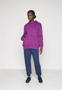 Sweat à capuche violet avec une poche avant et un cordon ; associé à un pantalon bleu marine et des chaussures de sport noires. Texture lisse partout.