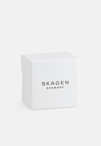 Emballage carré blanc aux bords arrondis, avec le texte "SKAGEN DANEMARK" dans une police simple et moderne. Texture lisse et mate.