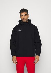 Czarna, kapturowa kurtka przeciwdeszczowa z pełnym zamkiem, elastycznymi mankietami oraz białym logo Adidas na górnej lewej stronie. Gładka tekstura i lekka konstrukcja.