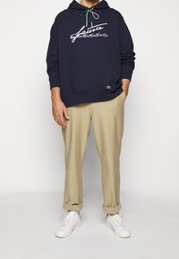 Námořnicky modrá mikina s kapucí a bílým textem, béžové kalhoty a bílé tenisky. Má přední kapsu a zelené detaily na lemu.