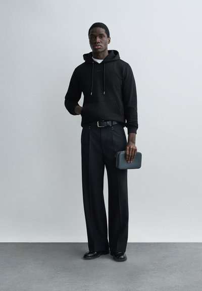 Άνδρας που φοράει μαύρο φούτερ με κουκούλα, μαύρο παντελόνι φαρδύ, μαύρα παπούτσια, κρατώντας μια μικρή μαύρη τσάντα χειρός, στέκεται μπροστά σε απλό γκρι φόντο.