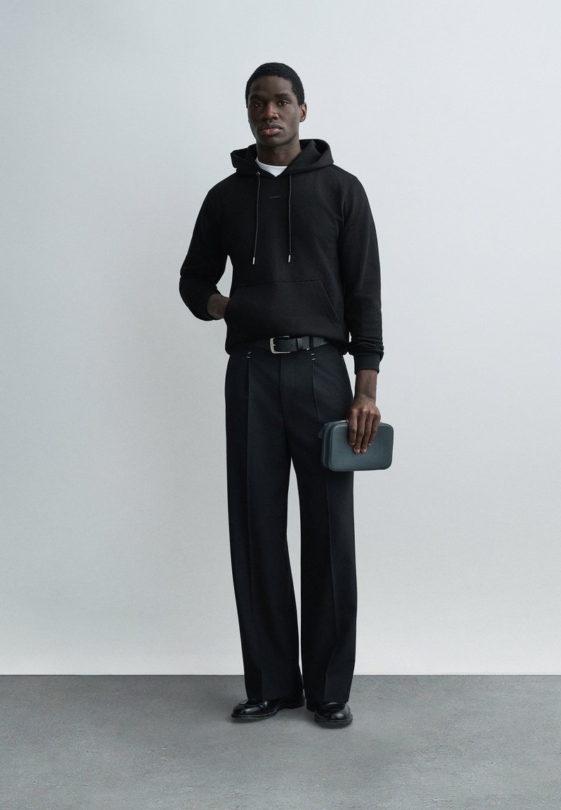 Mann trägt schwarzen Hoodie, schwarze weite Hosen, schwarze Schuhe, hält eine kleine schwarze Clutch-Tasche und steht vor einem schlichten grauen Hintergrund.