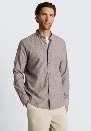 Homme portant une chemise marron clair à boutons avec col mandarin et un pantalon beige, debout devant un fond uni gris clair.