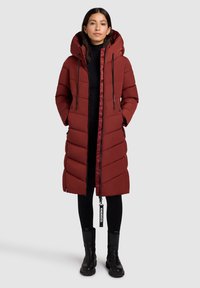Mujer vistiendo un abrigo de invierno largo y acolchado rojo con capucha, leggings negros y botas de tobillo negras, de pie contra un fondo sencillo.