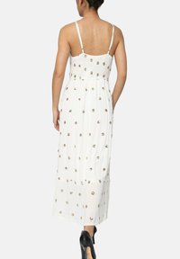 Robe maxi blanche avec bretelles spaghetti ajustables, tissu texturé et motifs floraux dorés ; ourlet en couches. Portée avec des talons noirs.