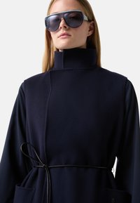 Navy fleece vest with high collar, black belt at the waist, side pockets, and layered long sleeves, paired with oversized sunglasses.