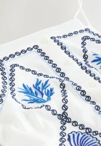 Bela bluzica iz tkanine z modrim in mornarsko modrim vezenjem, okrasnimi zavitki in gumbi. Vsebuje morsko-tematske vzorce.