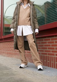 Lång olivgrön puffjacka över en ljusbrun v-ringad tröja och vit skjorta, i kombination med beige kordfläskbyxor och vita sneakers.