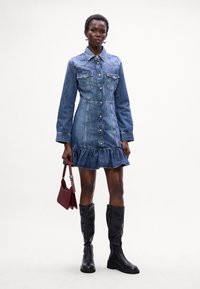 Vestido camisa de ganga com mangas longas, bolsos no peito, fecho de botão e bainha franzida. Combinado com botas pretas até ao joelho e uma pequena mala borgonha.