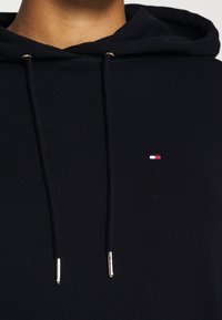 Zbliżenie na osobę ubraną w granatowy hoodie z małym logo w kolorach czerwonym, białym i niebieskim na klatce piersiowej oraz czarnymi sznurkami z metalowymi końcówkami.