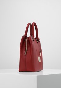 Röd läderhandväska med texturerad yta, dubbla topphandtag, guldfärgad hårdvara och en liten hängande etikett. Dragkedjestängning på ena sidan.