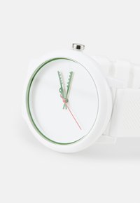 Reloj de pulsera blanco con una carátula redonda, acentos verdes y manecillas de horas y minutos en forma serrada. Hecho de plástico con una correa texturizada.