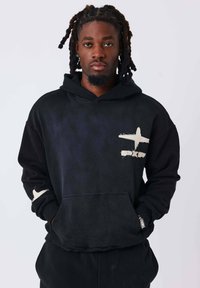 Sudadera negra con capucha que presenta un bolsillo frontal, tela texturizada y un gráfico de avión gris claro en el pecho izquierdo y en la manga.