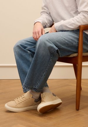 Beige leather sneakers with white soles, worn with light blue denim jeans featuring a relaxed fit and frayed hem, seated on a wooden chair.