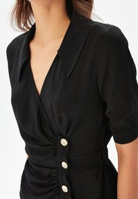 Robe noire avec un décolleté en V profond, manches courtes et détails froncés sur les côtés. Cinq boutons décoratifs de couleur claire ornent le devant.