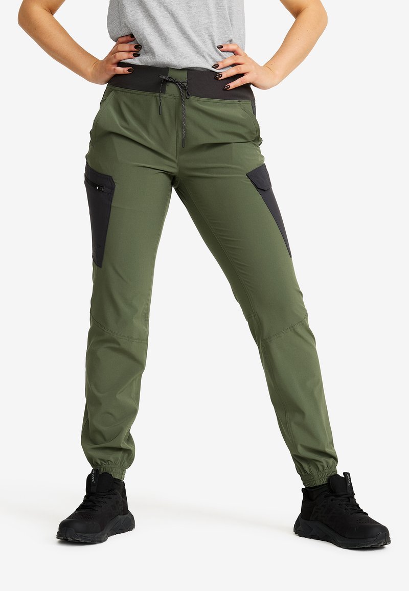 Person wearing olive green cargo pants with black side pockets, black waistband, black sneakers, standing with hands on hips.