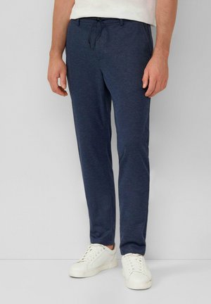 Mann trägt marineblaue, schlank geschnittene Freizeithose mit Kordelzugbund und weiße Turnschuhe, steht vor hellem, einfarbigem Hintergrund.