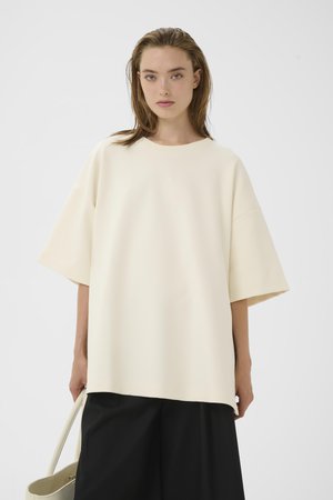 Kvinde med vådt hår iført oversized cremefarvet kortærmet top og sorte vidde bukser, der holder en cremefarvet håndtaske mod en ensfarvet baggrund.