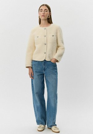 Veste bouffante couleur crème avec un col rond, ornée de boutons argentés et de deux poches avant, portée avec des jeans bleu clair.