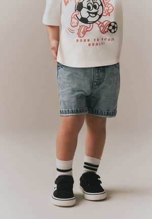 Enfant portant un t-shirt blanc à thème football, un short en jean, des chaussettes blanches à rayures noires et des baskets noires, debout sur un sol clair.
