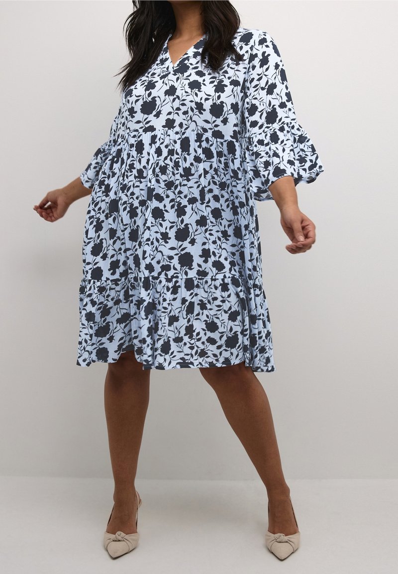 Femme portant une robe bleu clair mi-longue imprimée de fleurs noires avec des manches trois-quarts à volants, assortie à des talons pointus beiges.