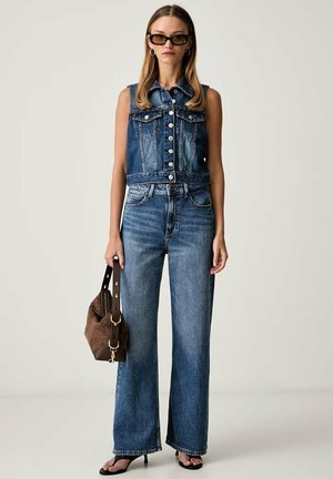Donna che indossa un gilet in denim senza maniche, jeans a vita alta con gamba larga, occhiali da sole neri, orecchini a cerchio, sandali neri con tacco, tiene una borsa marrone.
