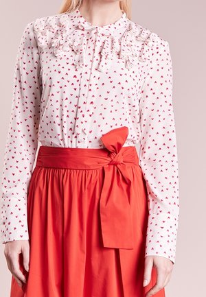 Mujer con blusa blanca con pequeños estampados de corazones rojos y canesú con volantes, combinada con una falda roja de talle alto atada con un lazo en la cintura.