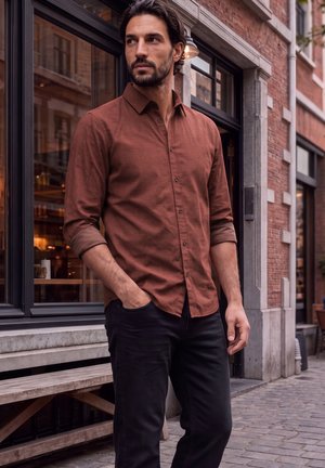 Un homme barbu, vêtu d’une chemise boutonnée couleur rouille et d’un pantalon noir, se tient devant une boutique en briques et regarde sur le côté.