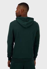 Sudadera verde oscura con una textura suave, que presenta una capucha con cordón, puños acanalados y un corte relajado. Sin logotipos ni patrones visibles.