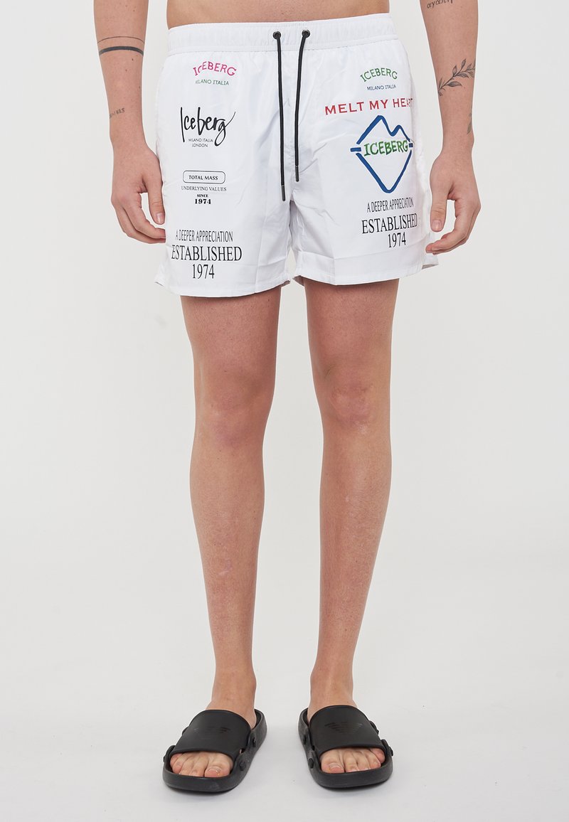 Pantaloni da bagno bianchi realizzati in un tessuto leggero con cordino nero, con loghi e testo stampati colorati sul fronte. Infradito nere.