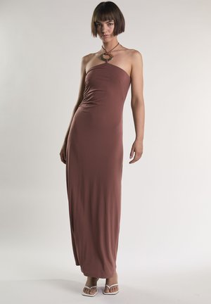 Femme portant une robe marron sans bretelles, longue jusqu'au sol, avec un détail en anneau métallique au niveau de l'encolure et des talons blancs ouverts aux orteils, debout sur un fond uni.