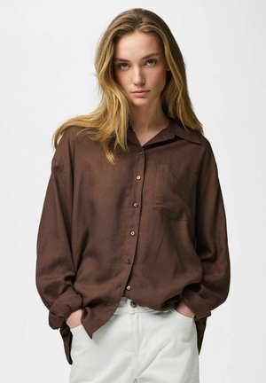 Fiatal nő szőke hajjal, laza barna gombos ingben és fehér nadrágban, kezét zsebébe dugva áll egy egyszínű háttér előtt.