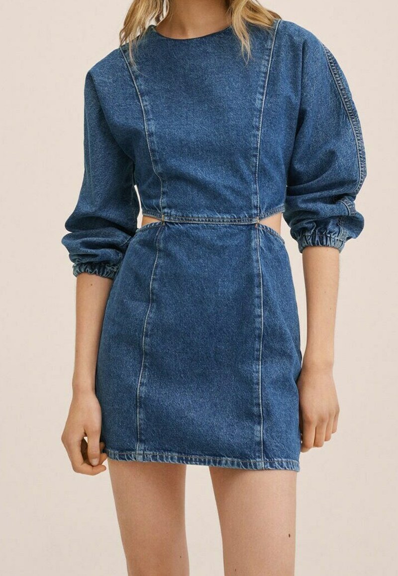 Femme portant une mini robe en jean bleu avec des manches longues bouffantes et des découpes sur les côtés à la taille, sur un fond uni.
