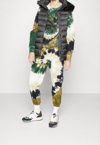 Svart pufferväst med fuskpäls på huvan, grön och brun tie-dye tröja, matchande joggers och flerfärgade träningsskor.