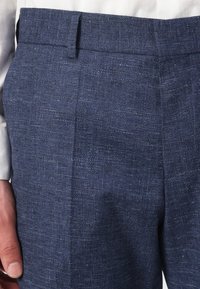 Közeli kép tengerészkék texturált öltöny nadrágról, fehér inggel viselve, megmutatva a derékpánt részleteit és az oldalsó varrást.