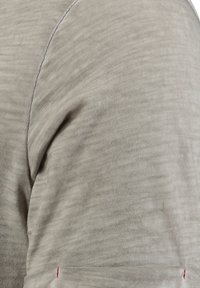 Camisa de manga corta de color beige hecha de un tejido texturizado. Presenta líneas horizontales sutiles y un pequeño acento rojo cerca del puño.