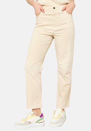 Femme portant un pantalon beige clair à jambes droites, les mains dans les poches, et des baskets blanches avec des touches jaunes, vertes et violettes.