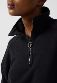Pull noir à col haut, avec fermeture éclair half-zip à l'avant et détail de fermeture éclair texturé, fabriqué en tissu doux et lisse.