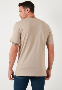 Tricou bej cu mânecă scurtă, realizat dintr-un material moale. Are un guler rotund și o textură netedă, cu tatuaje vizibile pe braț.