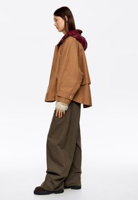Brązowa kurtka z bordowym kapturem, w oversize'owym kroju, z dużymi bocznymi kieszeniami i futrzanymi akcentami na rękawach. Połączona z luźnymi, zielonymi spodniami i ciemnymi butami.
