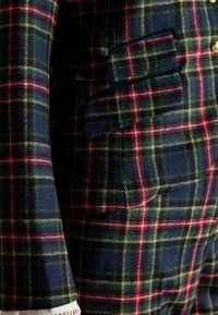 Wool blazer featuring a navy, green, and red tartan pattern. Includes a side pocket and white trim at the cuff for added detail.